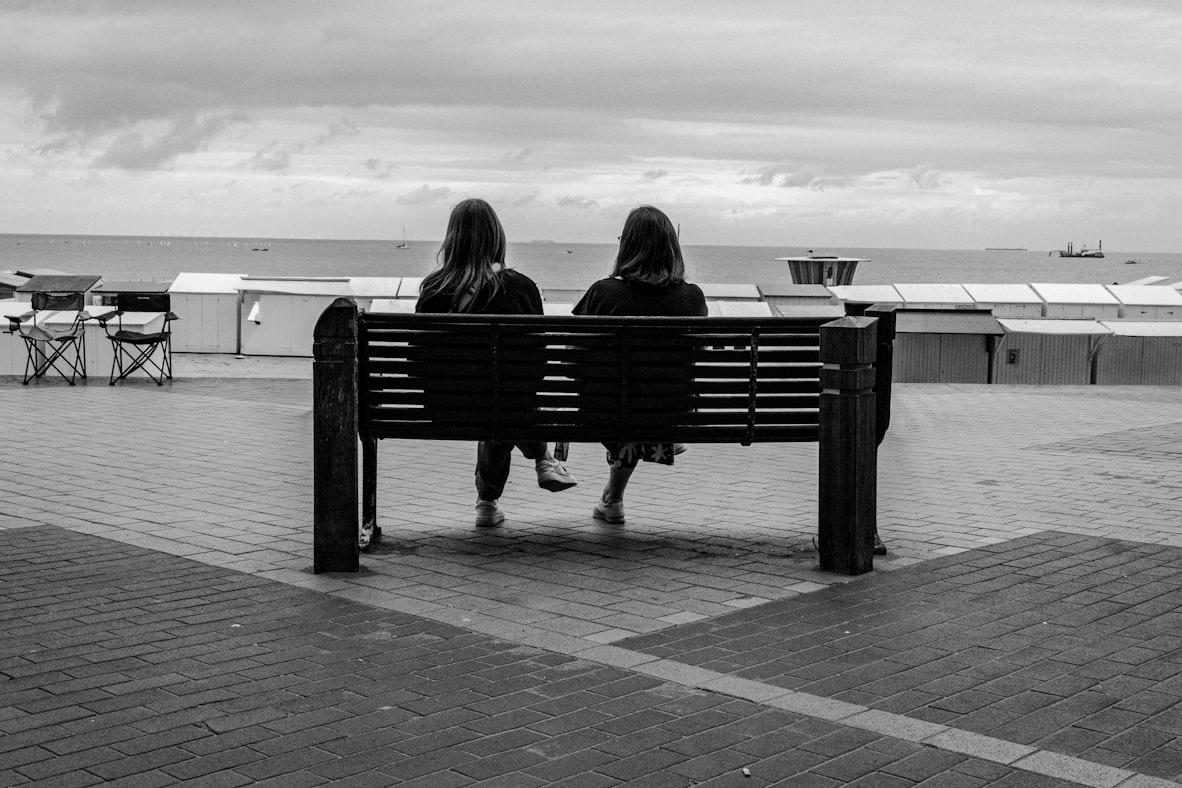 Two travelers sitting on a bench overlooking the ocean at golden hour, mid-conversation, neither one rushing to leave
