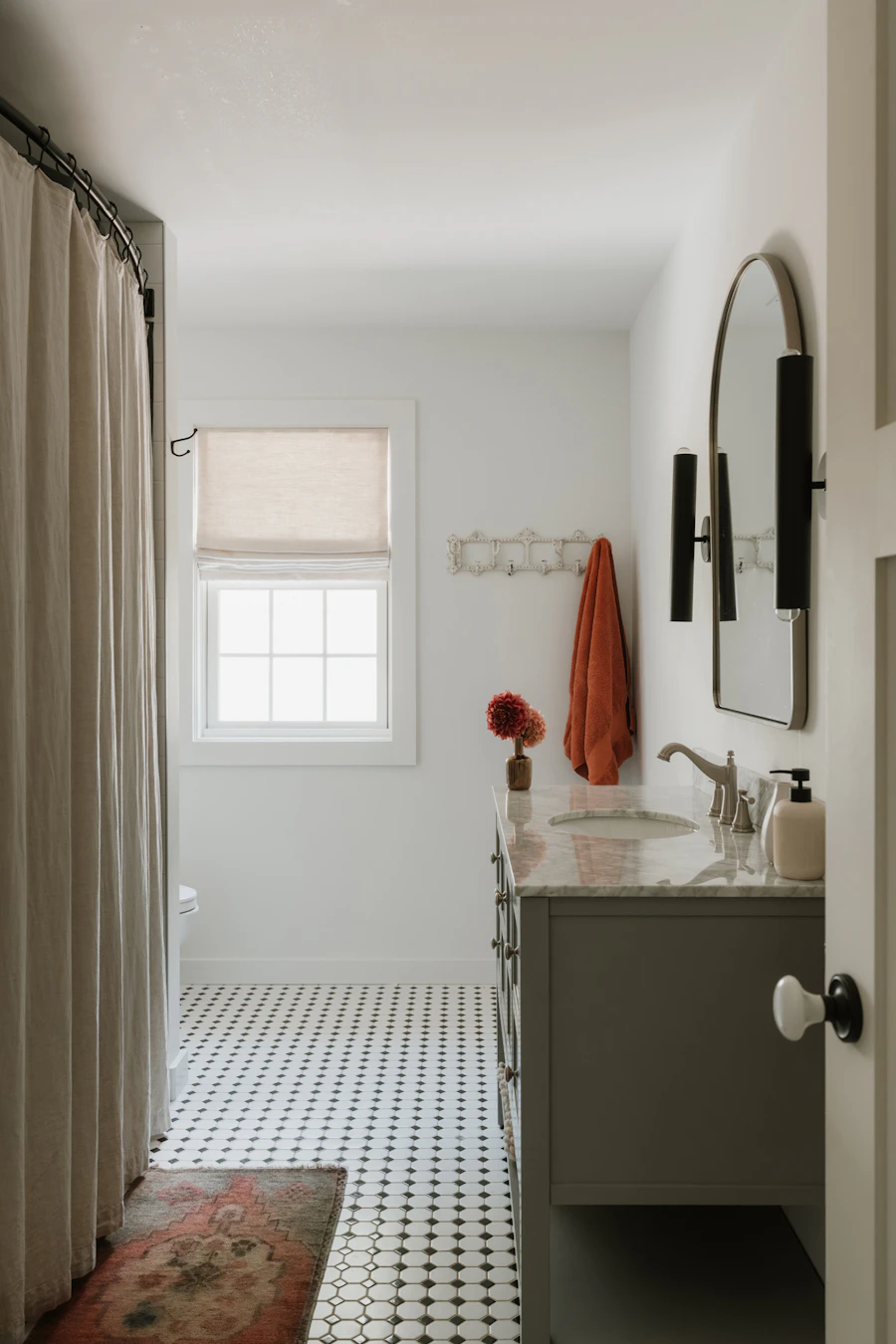 A compact bathroom with a vanity, mirror, wall lighting, shower curtain, and patterned tile floor.