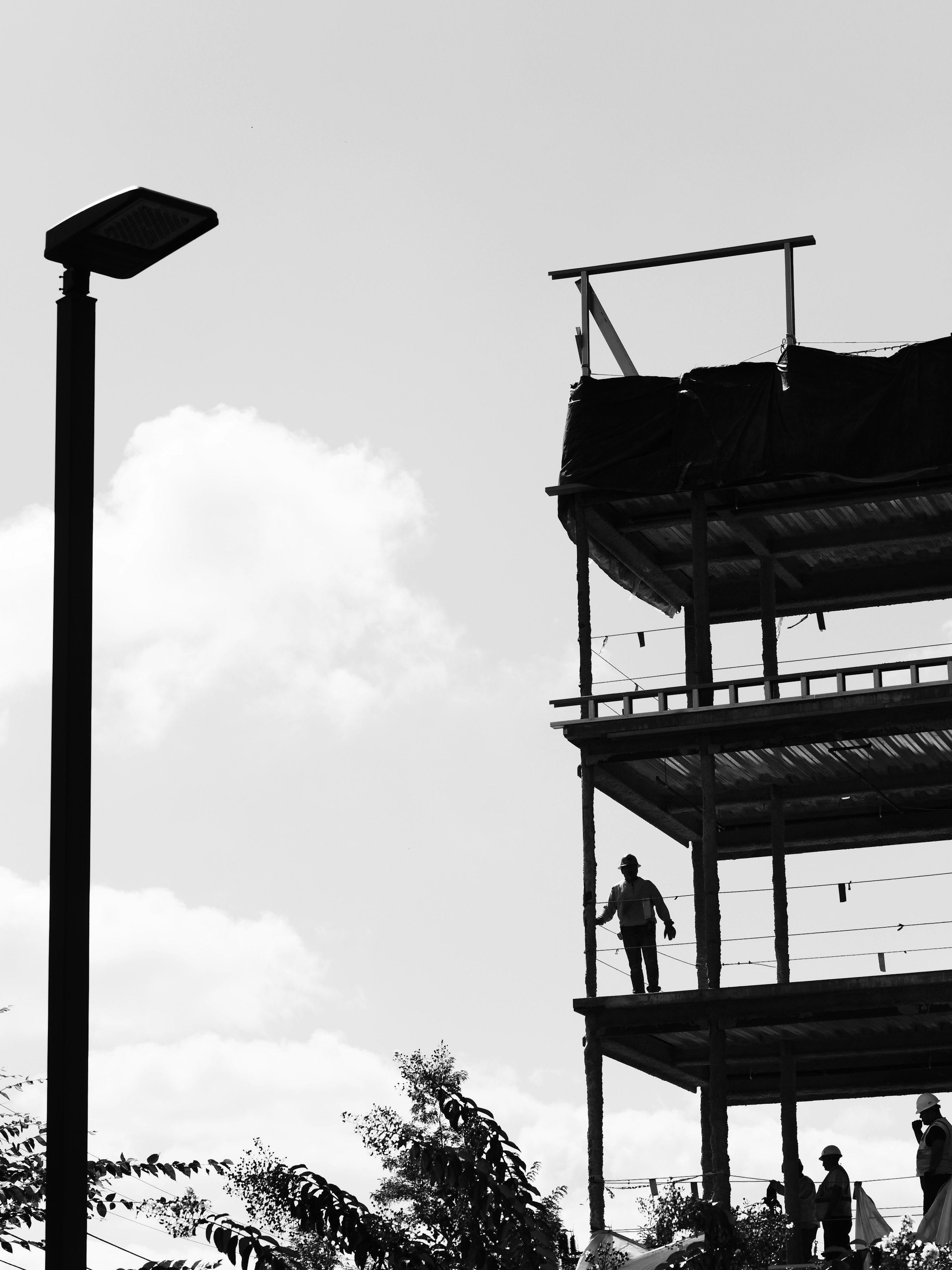 Construction workers moving along the open steel frame of an unfinished building, silhouetted against a pale sky.