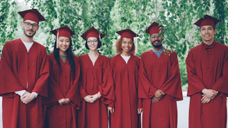 Six graduates in red gowns and caps stand together.