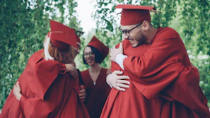 Graduates in red gowns hug each other happily.