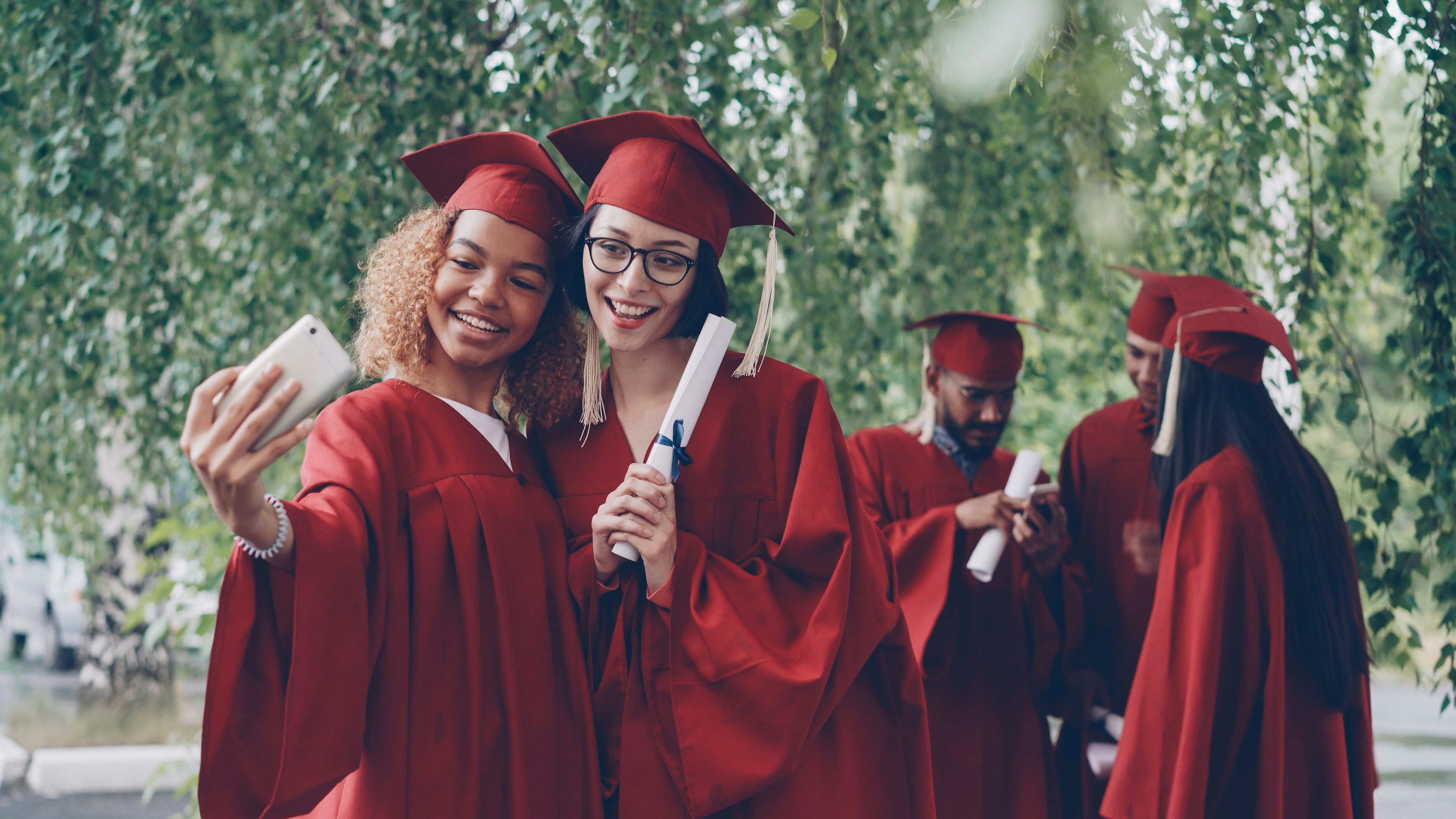 Graduates in red gowns taking a selfie with diplomas.