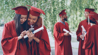 Graduates in red gowns look at a phone together.