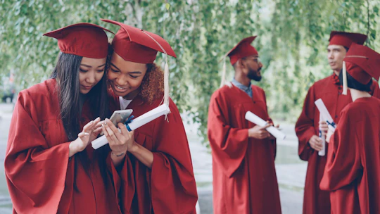Graduates in red gowns look at a phone together.