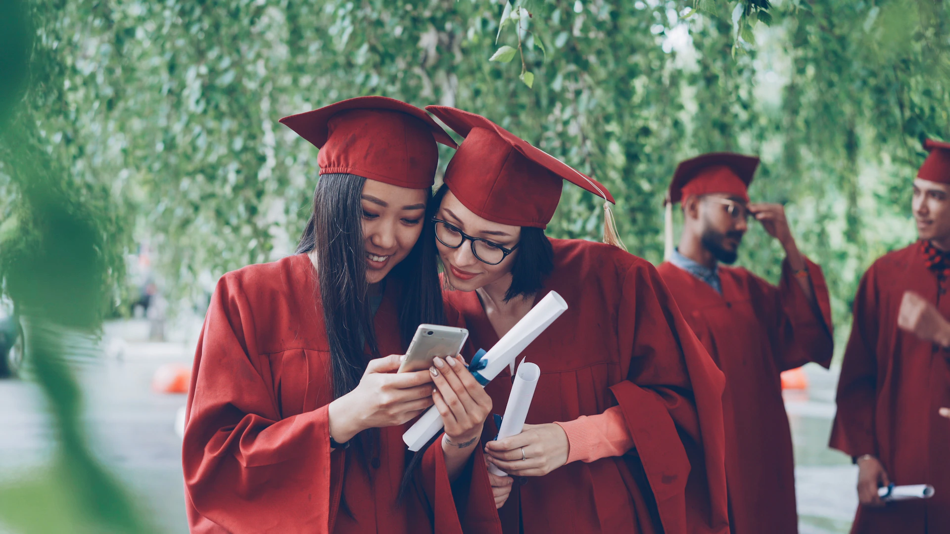 Two graduates in red gowns looking at a smartphone