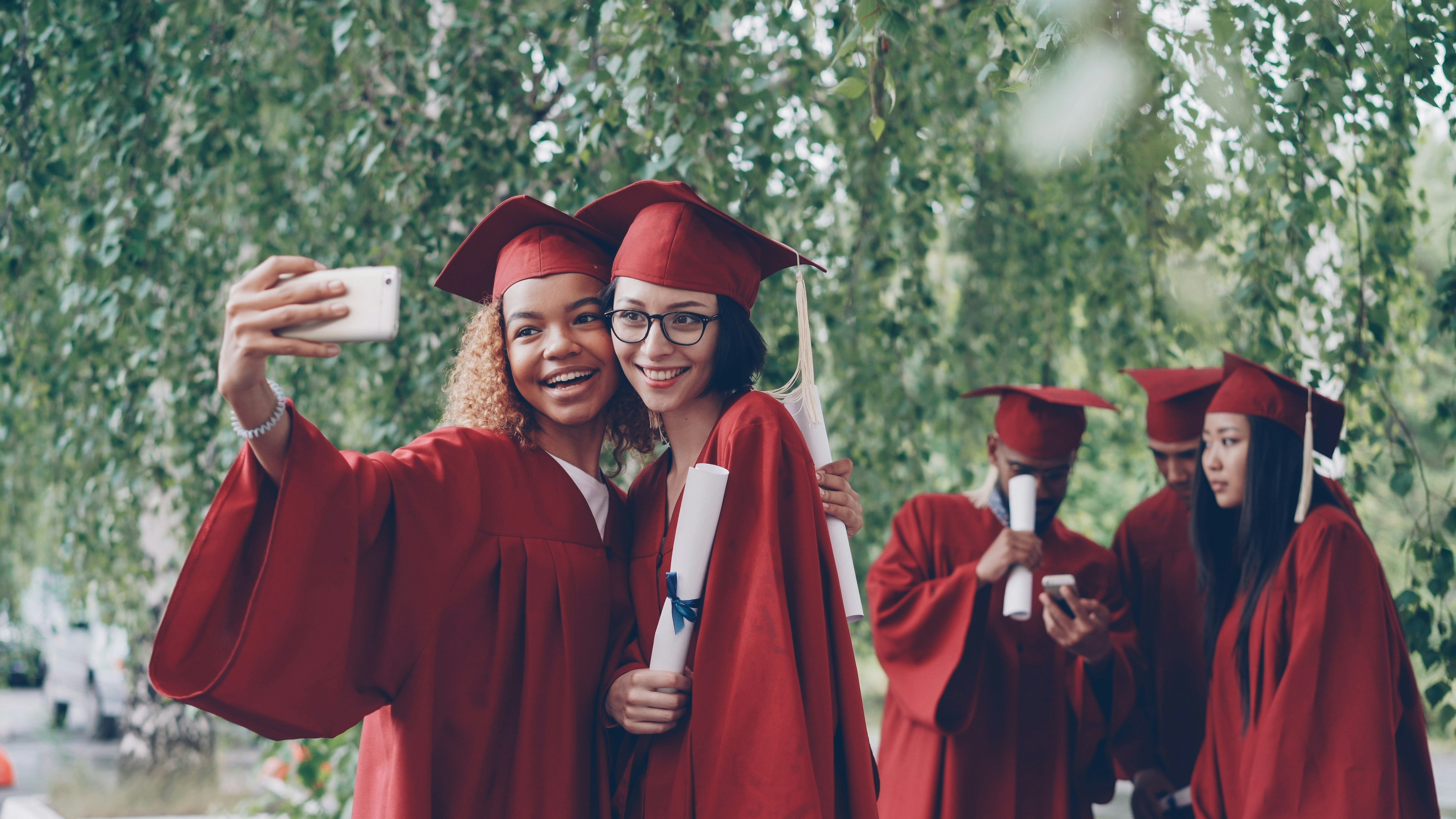 Two graduates in red gowns take a selfie with diplomas.