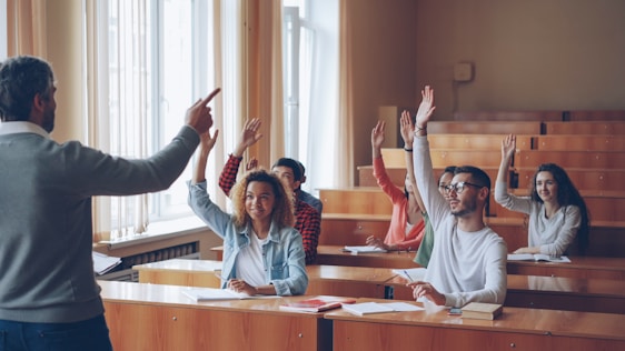 Teacher pointing at students with raised hands in lecture hall.