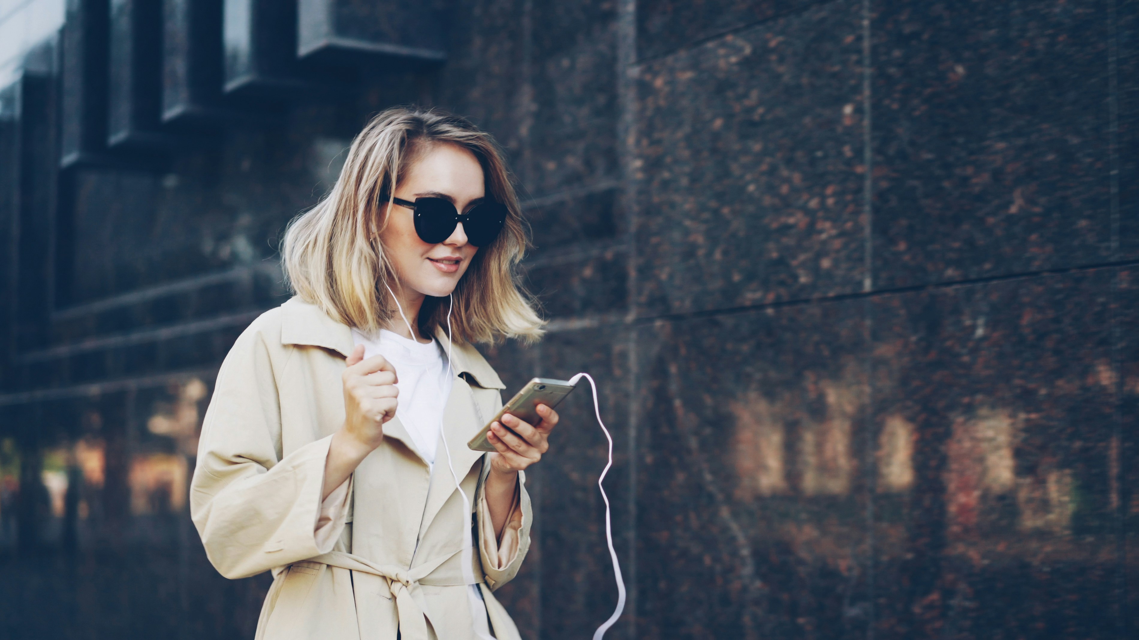 Pretty girl is listening to music through earphones using smartphone and dancing walking in the street and enjoying rhythm and modern city. Girl is wearing sunglasses and trendy coat.