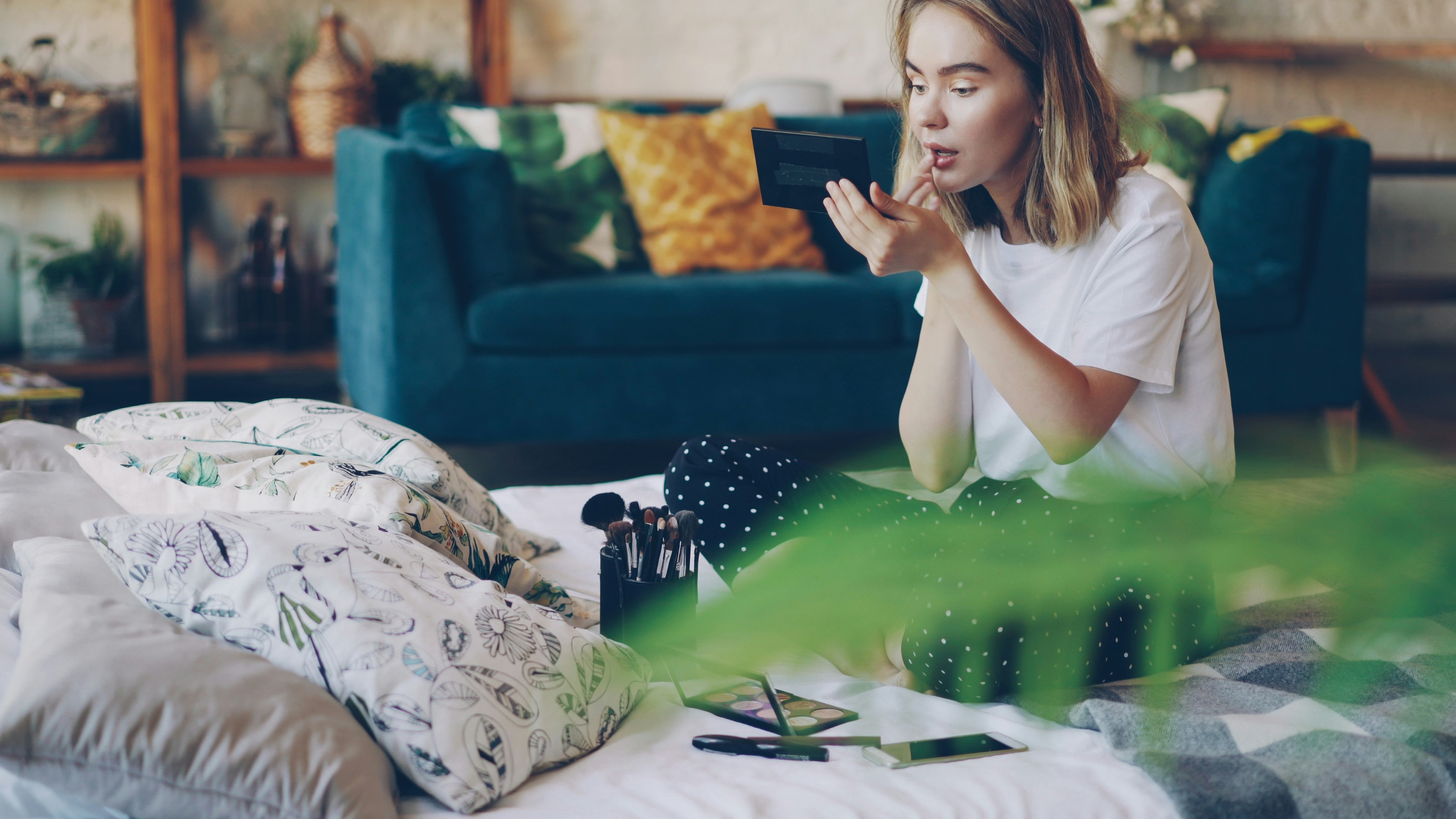 Beautiful blonde is painting lips putting on make-up using professional decorative cosmetics sitting on bed at home. Beauty, modern interiors and young women concept.