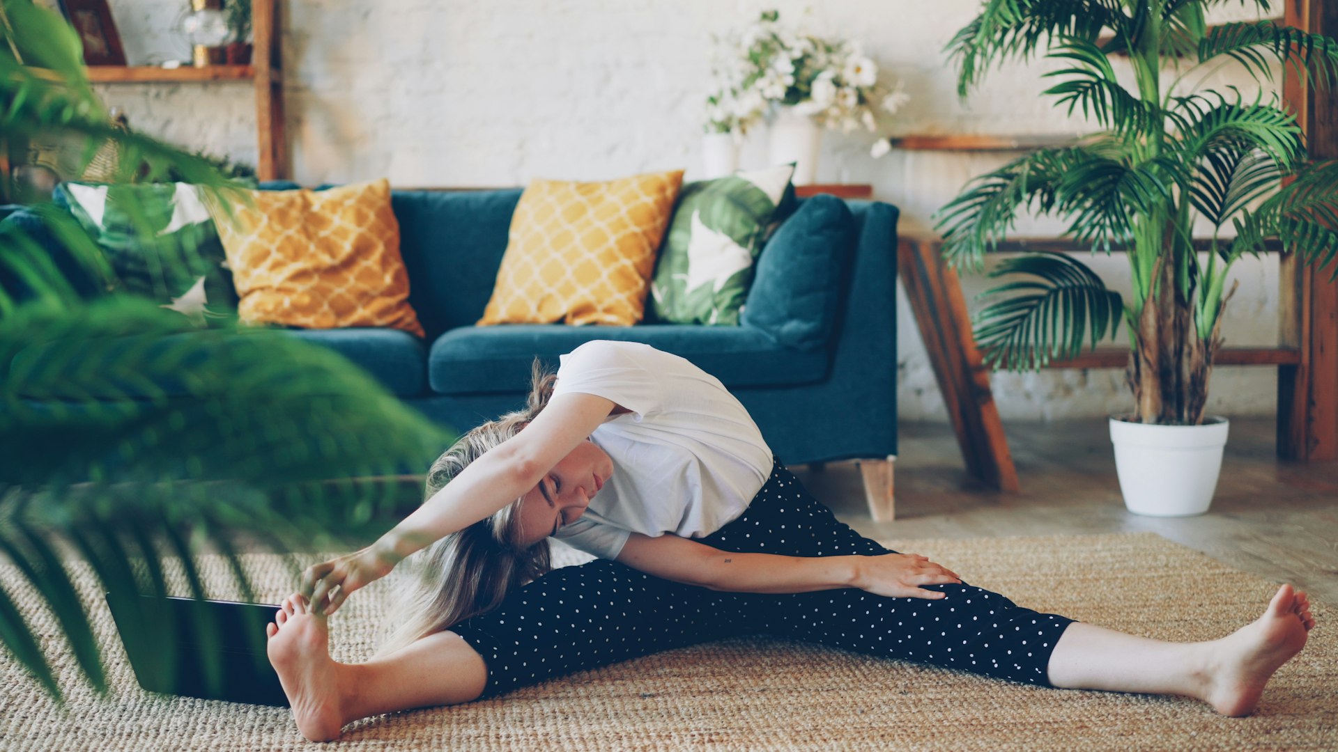 Woman sitting on a living room rug with her legs wide apart, reaching over one leg toward her foot
