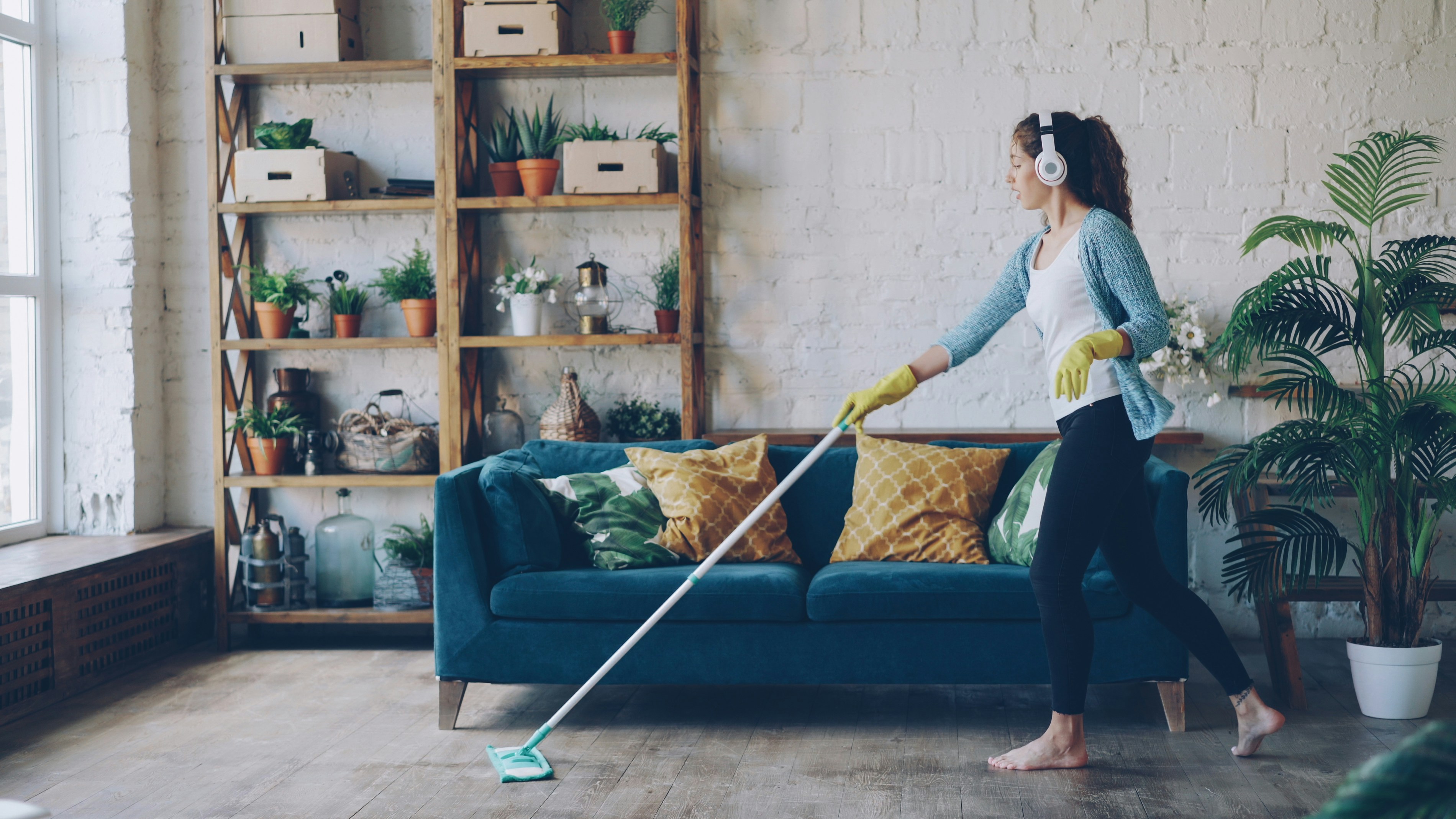 Cheerful young woman is dancing with mop during routine clean-up in nice loft style apartment, she is listening to music with wireless headphones and singing enjoying her occupation.