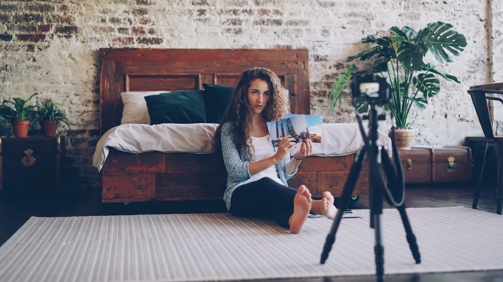 A creator sitting on the floor of a bright bedroom showing printed photos to a camera on a tripod
