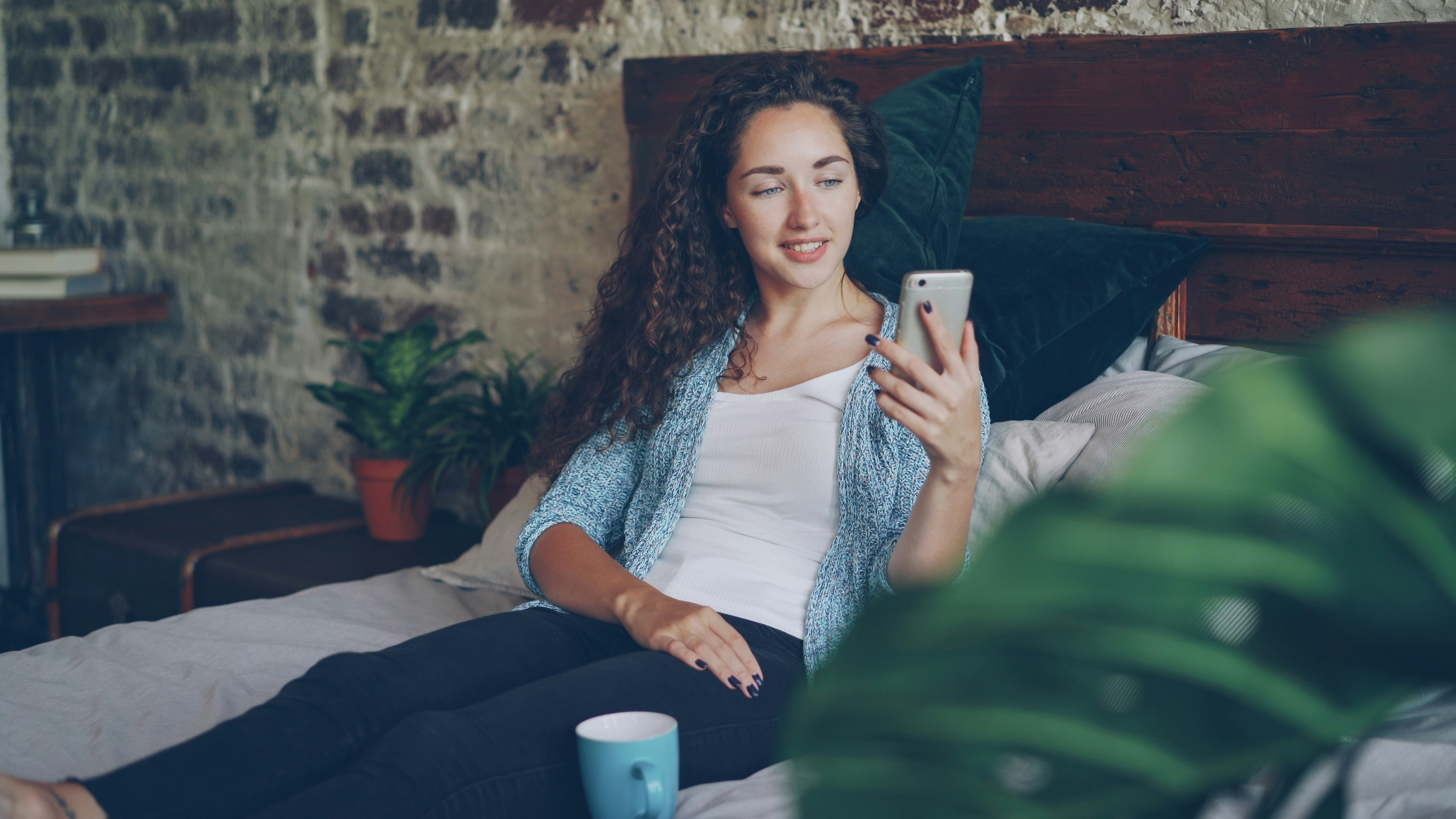 Good-looking woman is talking to friend online using modern smartphone making video call, she is happy and excited. Young lady is sitting on bed at home wearing casual clothes.