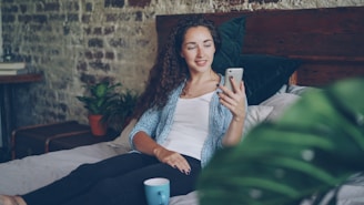 Young woman relaxing in bed with smartphone and coffee.