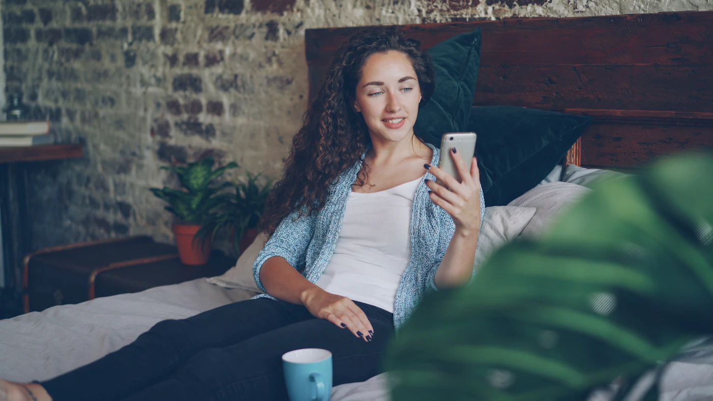 Mulher sorrindo ao olhar o celular, recostada na cama, com uma xícara ao lado — acompanhamento do ciclo menstrual