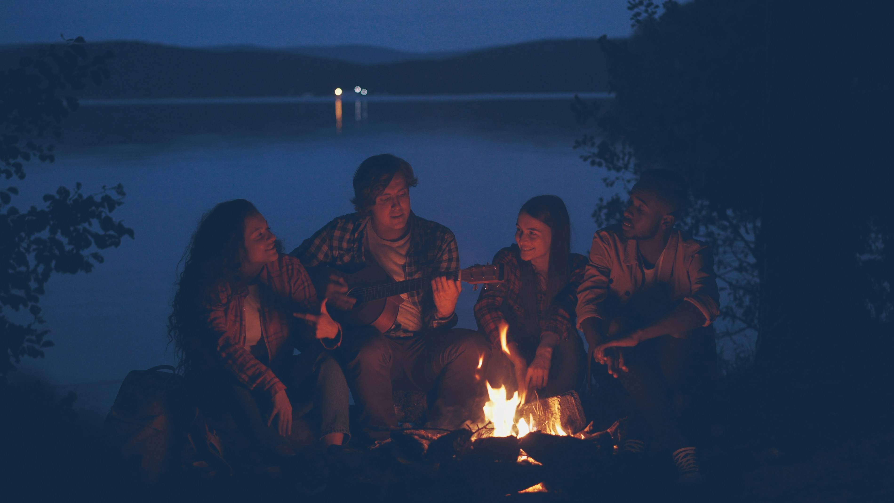 Friends gather around a campfire by a lake at dusk