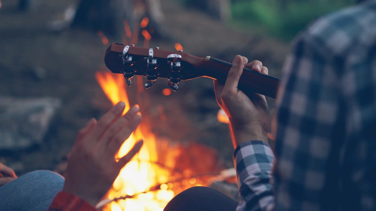 At night, hands playing an acoustic guitar around a campfire, silhouettes of people spending time around it.