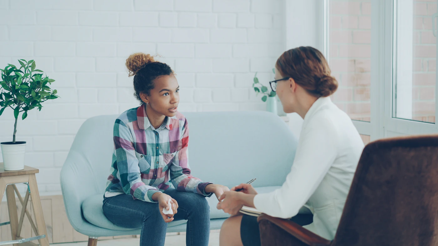 A counselor taking notes while a young woman speaks during a calm private session on a light couch