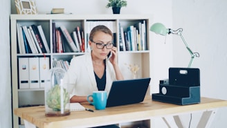 Woman on phone at desk with laptop and books.