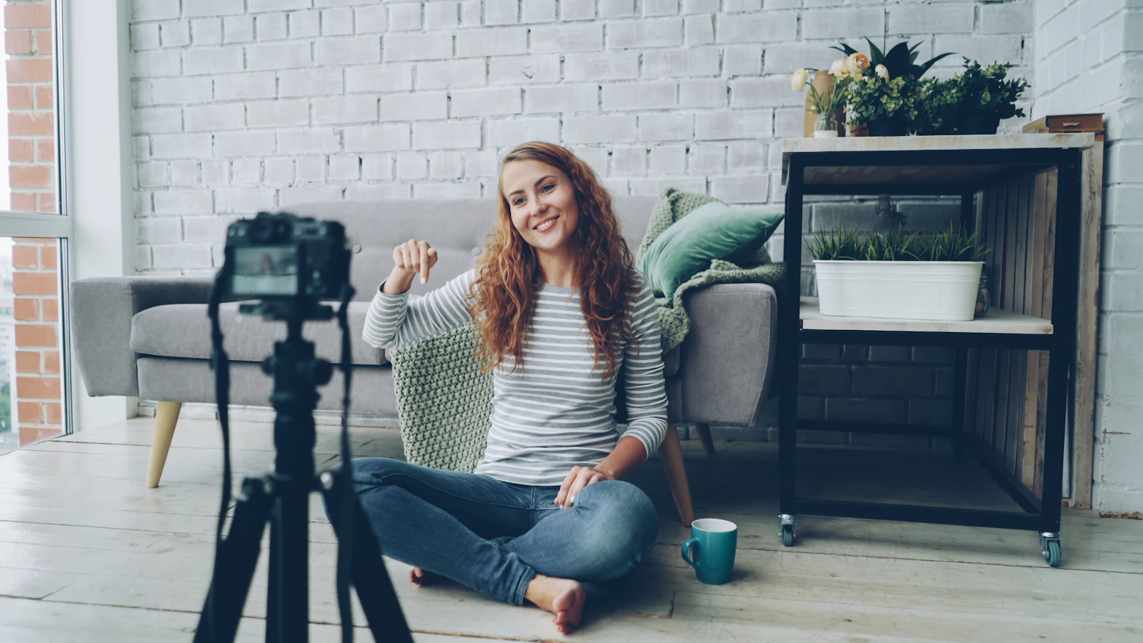 A woman sitting on the floor of a bright living room waving at a camera on a tripod while recording a video