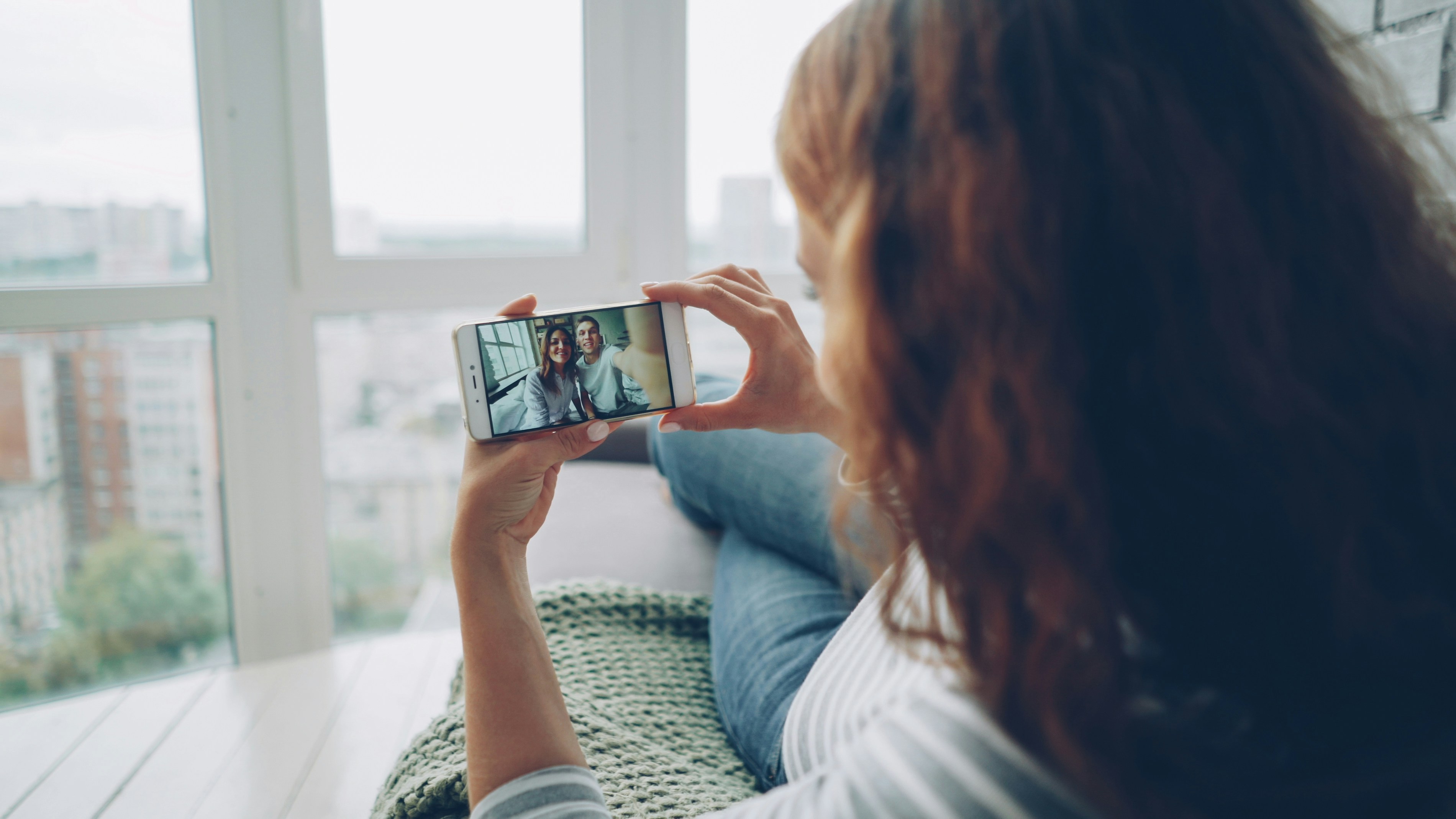 Cheerful young woman is chatting with friends using smartphone looking at screen and watching beautiful couple then showing thumbs up and waving hand. Modern communication concept.