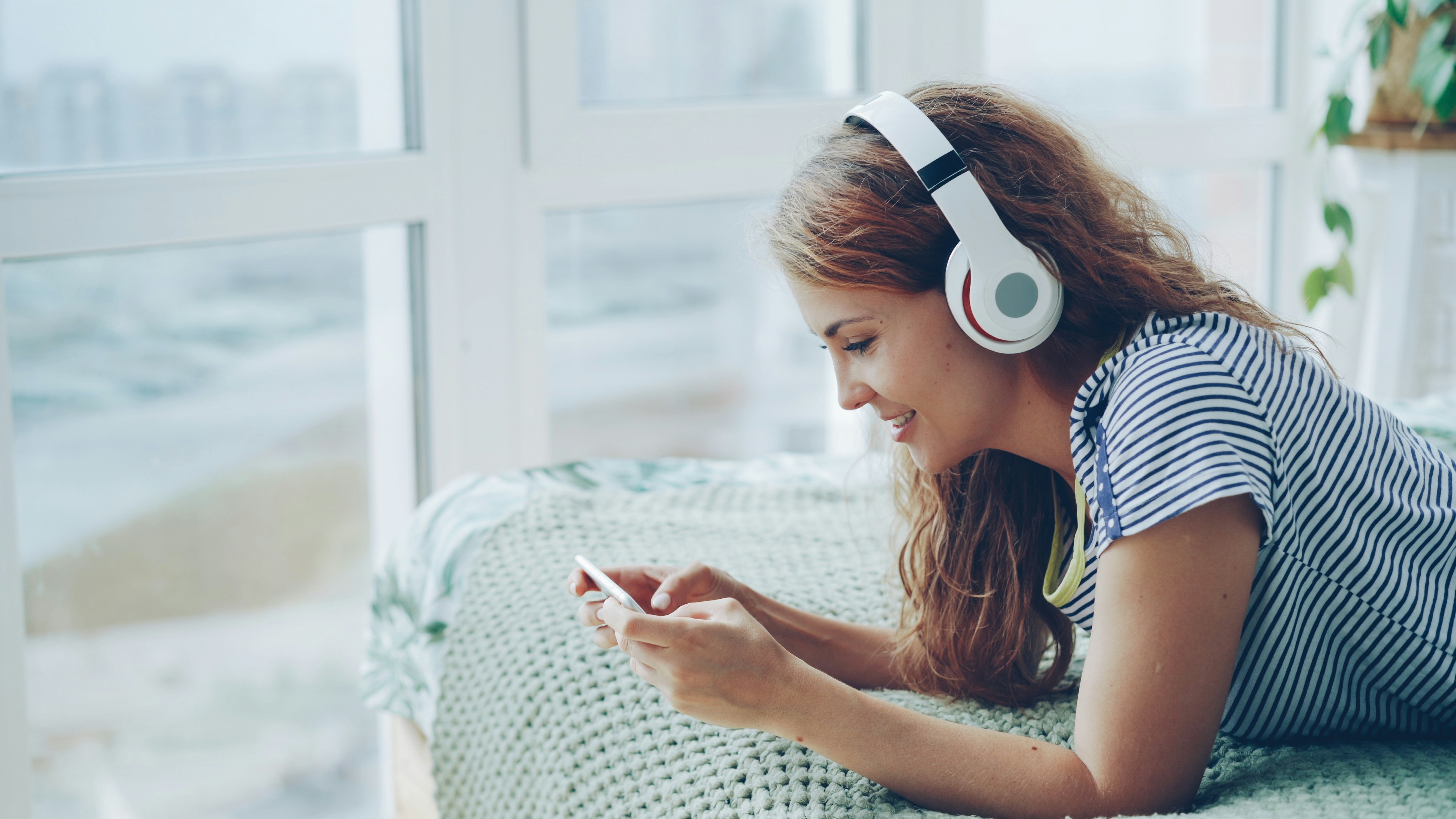 Young woman wearing headphones and using smartphone on bed.