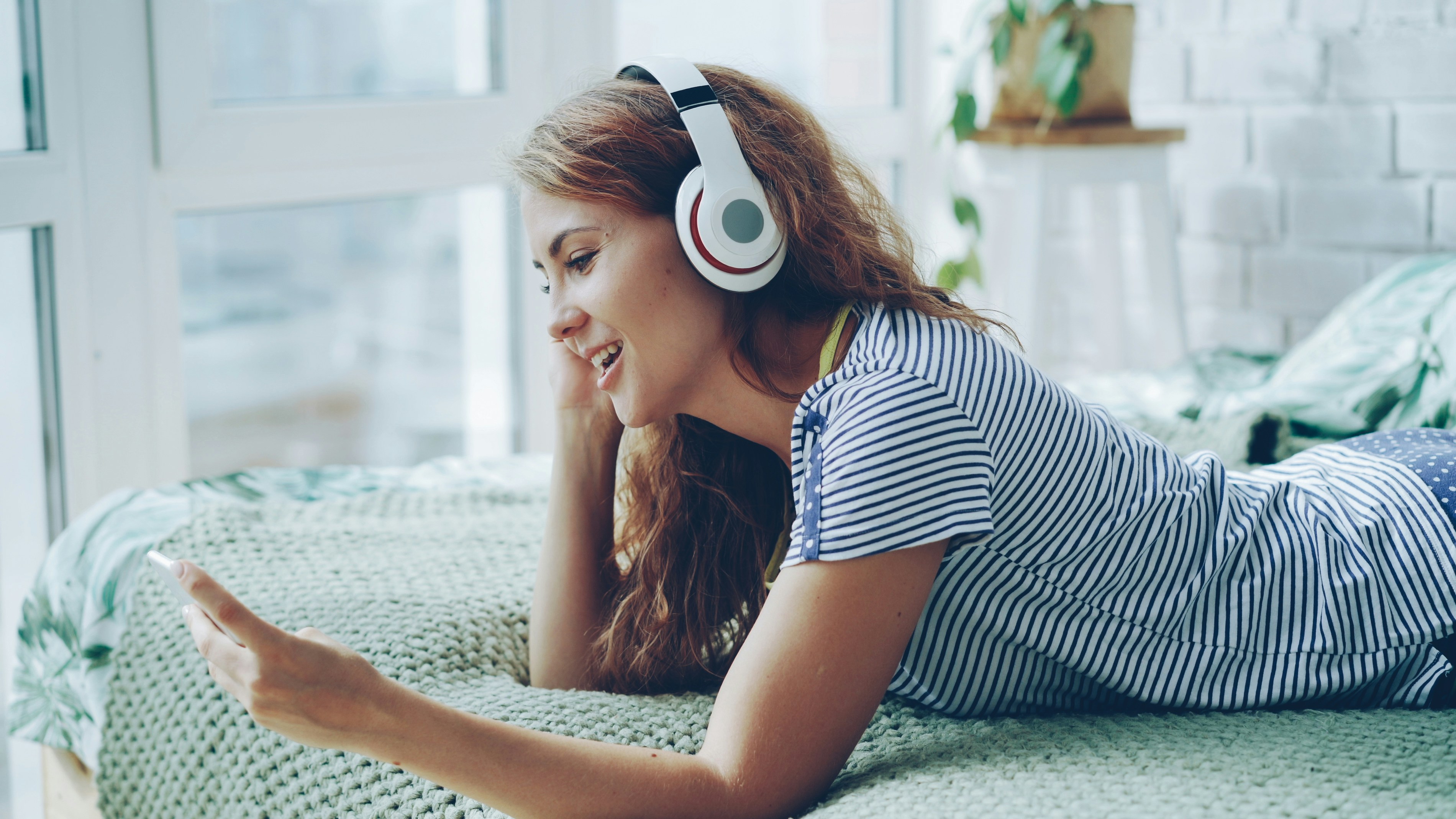 Young woman wearing headphones and using phone on bed.