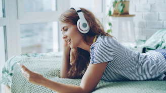 Young woman wearing headphones and using phone on bed.