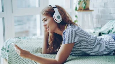 Young woman wearing headphones and using phone on bed.