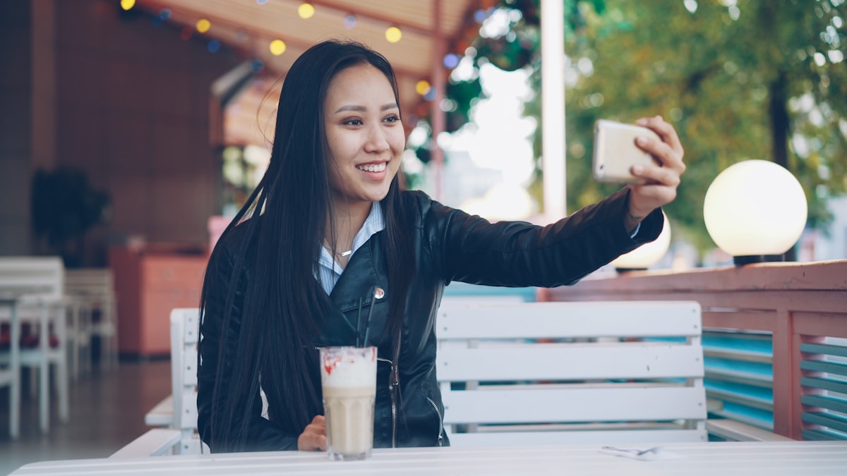 Asian woman taking a selfie at an outdoor cafe with a latte on the table
