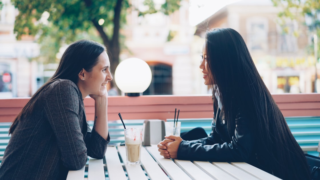 Two people sitting at an outdoor cafe table talking to each other, representing shoppers routing around AI recommendations to reach human opinions before buying