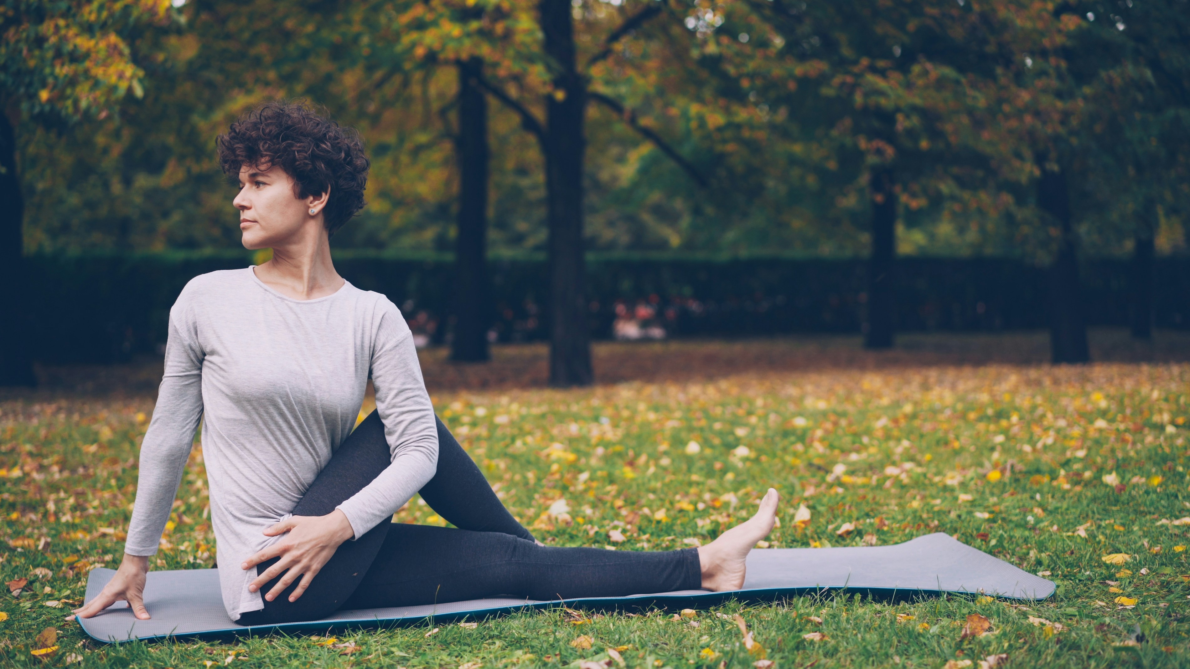 Slim young lady is doing sequence of yoga asanas sitting on mat in park alone on warm autumn day with beautiful grass and trees around her. Recreation and sport concept.