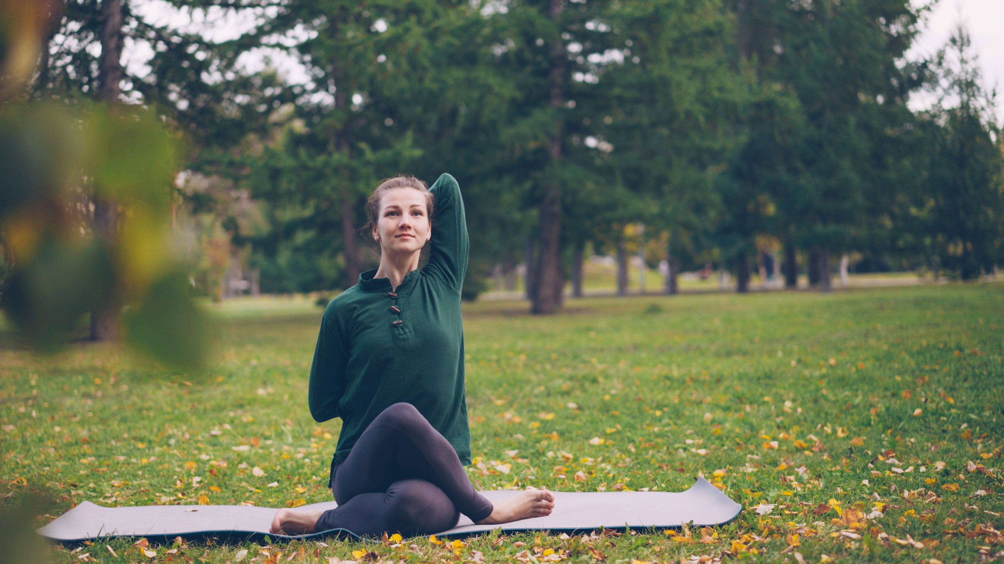 Cheerful young lady is exercising in park sitting in Cow Face pose with arms behind her back during outdoor practice in city park. Nature, millennials and health concept.