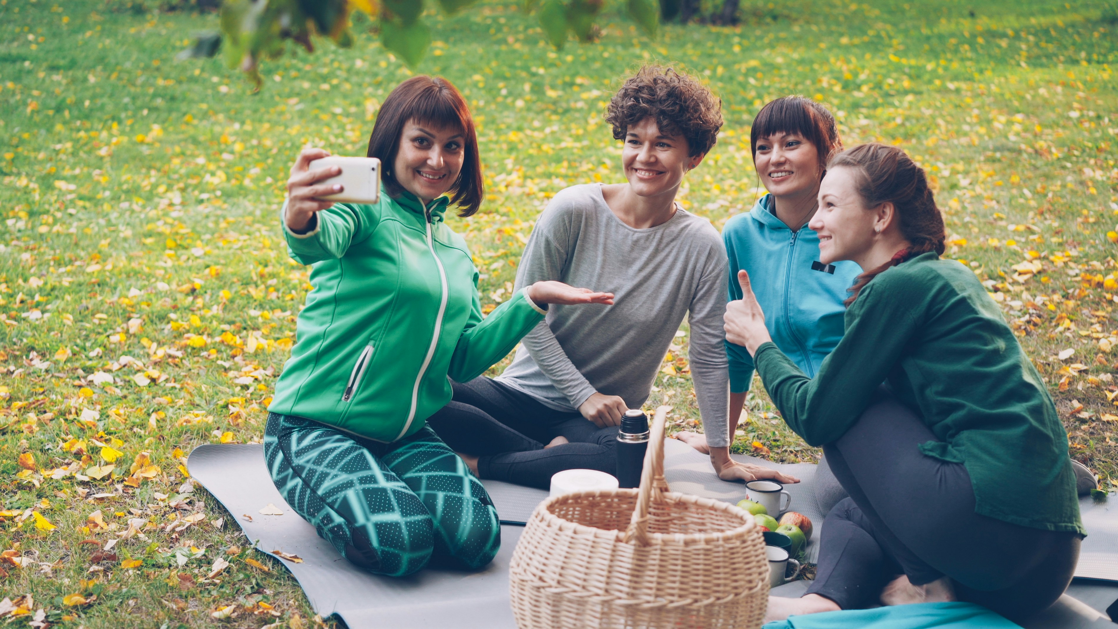 Pretty young women are taking selfie with smartphone during picnic in park sitting on grass, posing and laughing having fun. Nature, friendship and modern technology concept