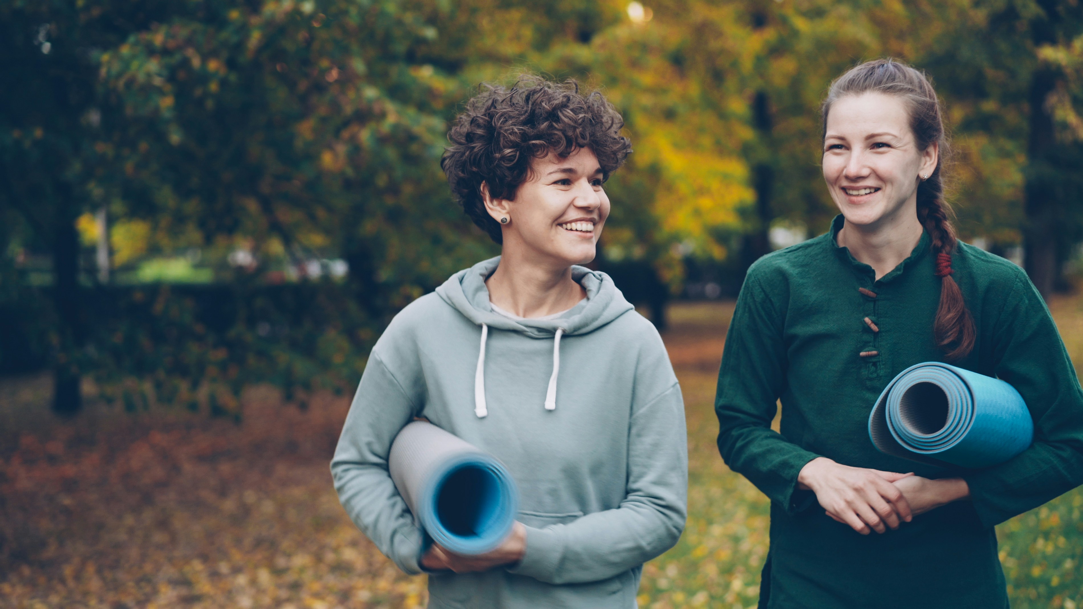 Two women holding yoga mats walking in a park.