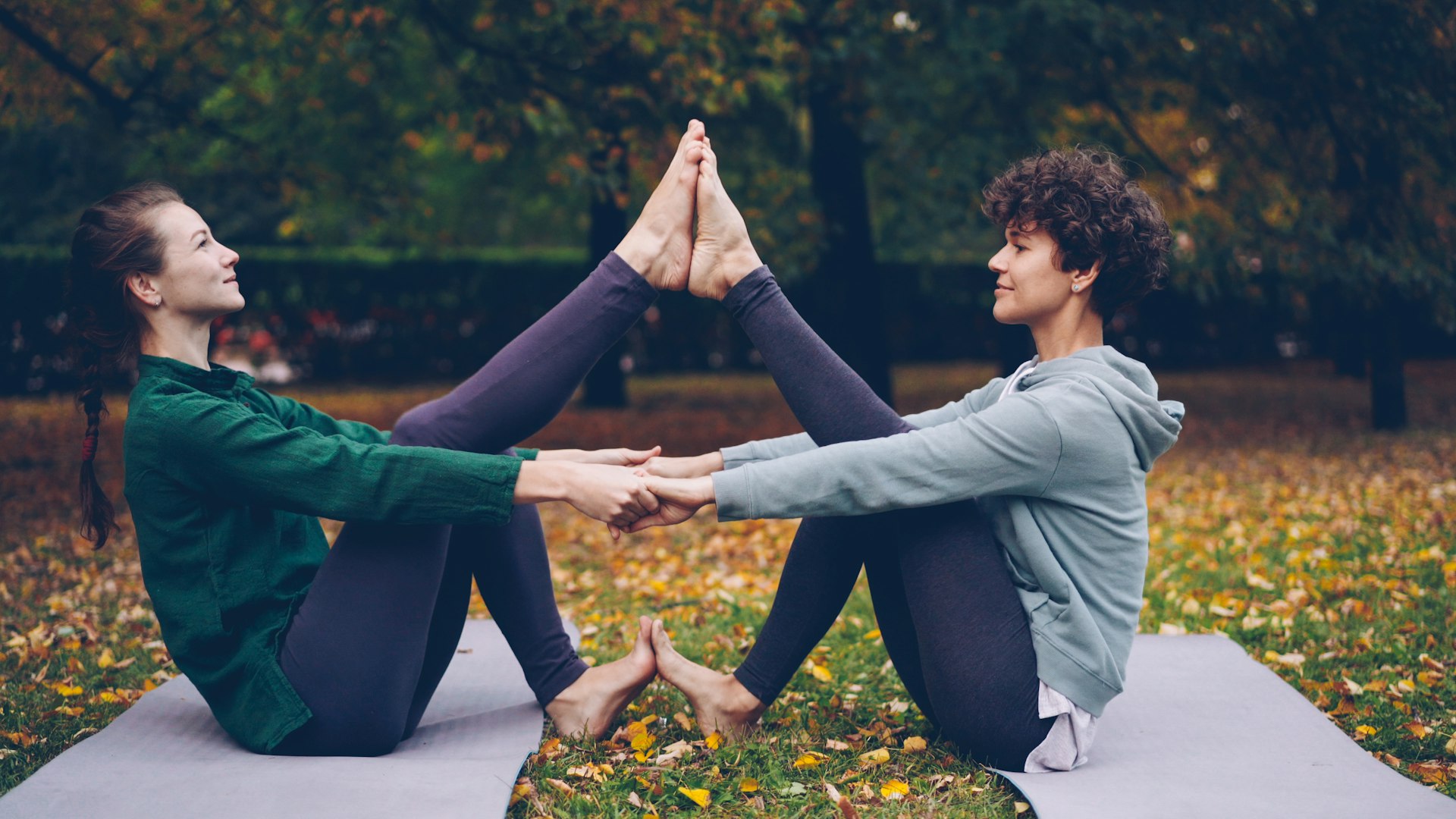 Two women sitting face to face on yoga mats in a park, holding hands with the soles of their feet pressed together