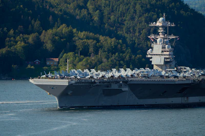Aircraft carrier sailing on water near forested hills.