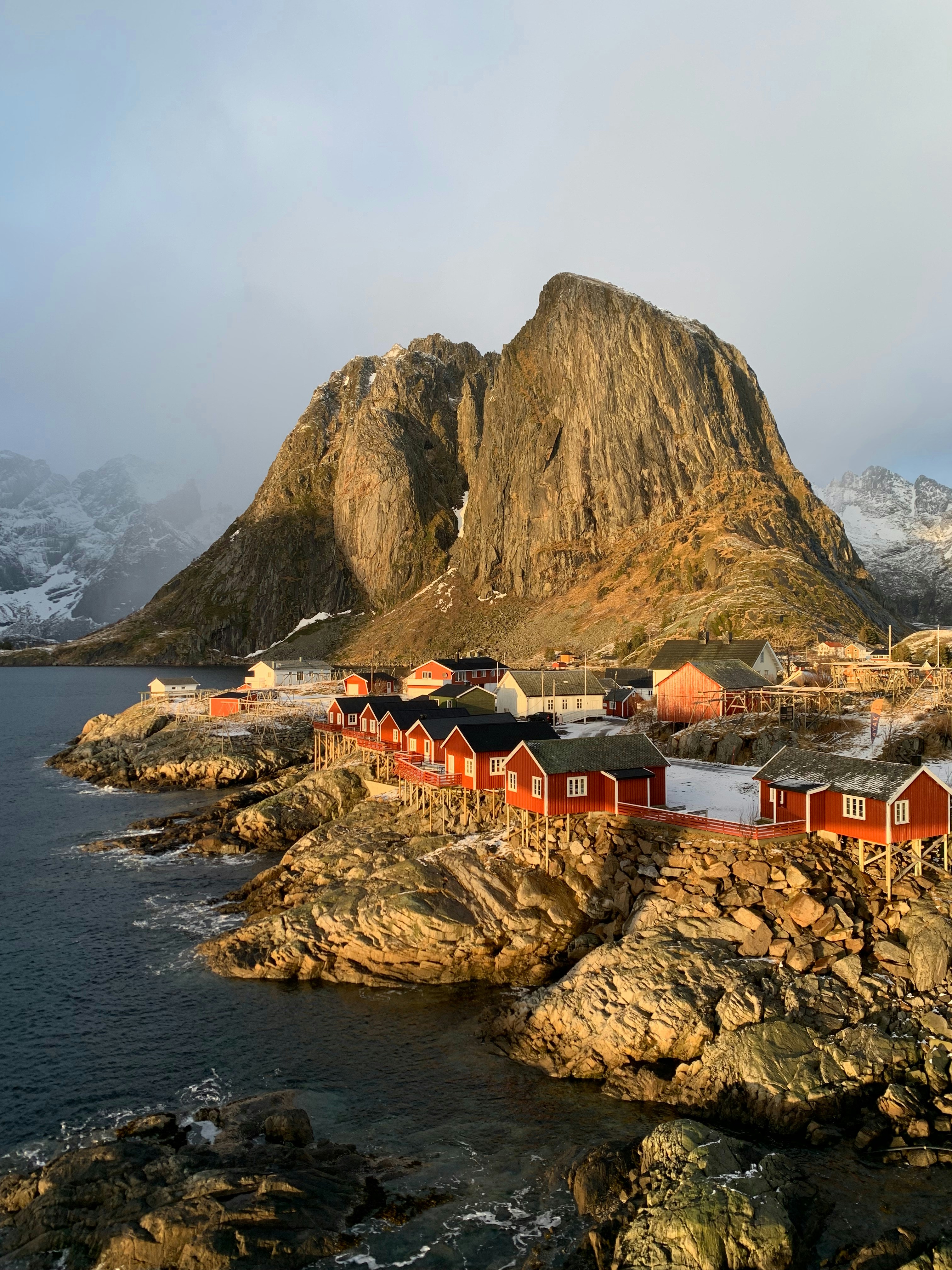 Red fishing cabins on rocky coast with mountain backdrop.