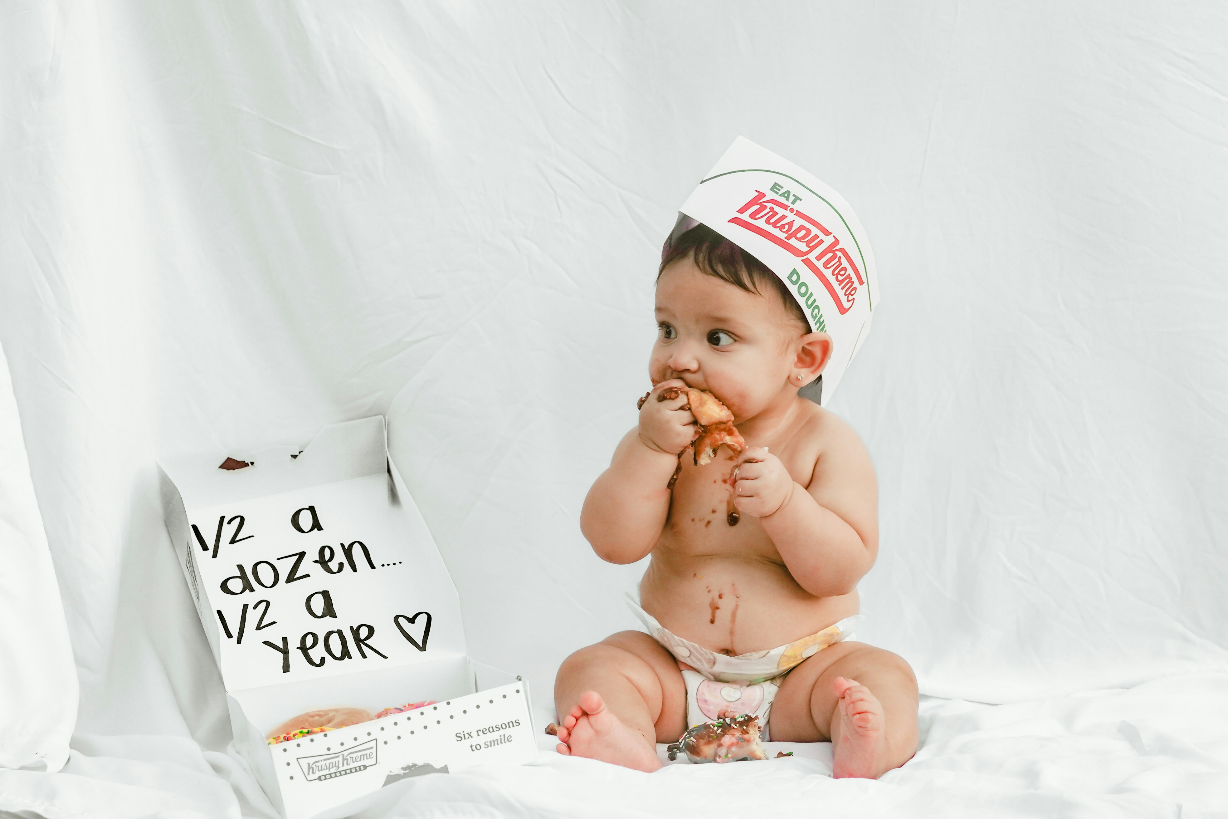 Baby wearing a krispy kreme hat eating a donut.