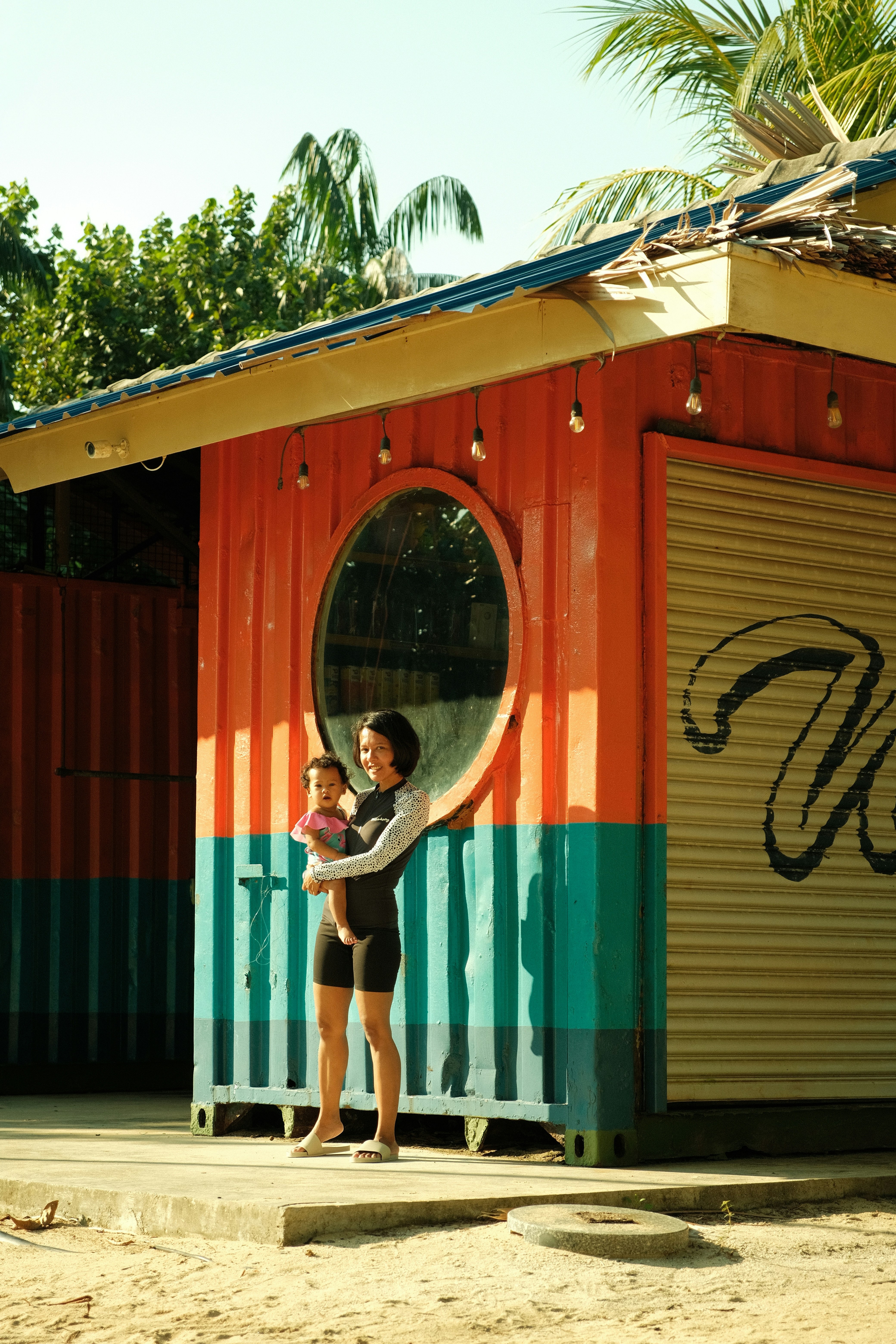 Femme tenant bébé devant un kiosque de plage coloré.