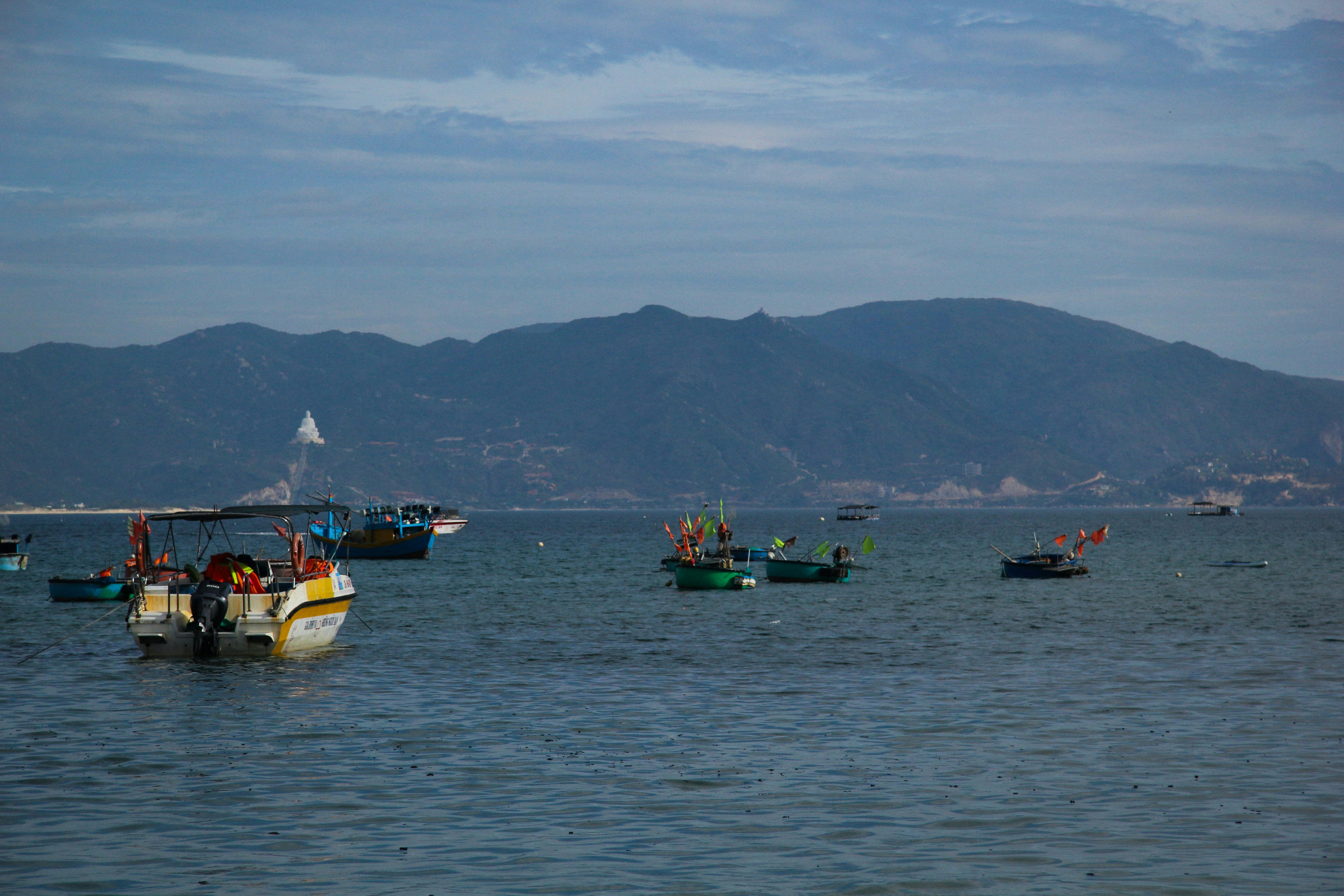 Fishing boats float on the calm sea with mountains behind.