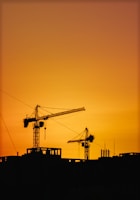 Construction cranes silhouetted against an orange sunset sky.