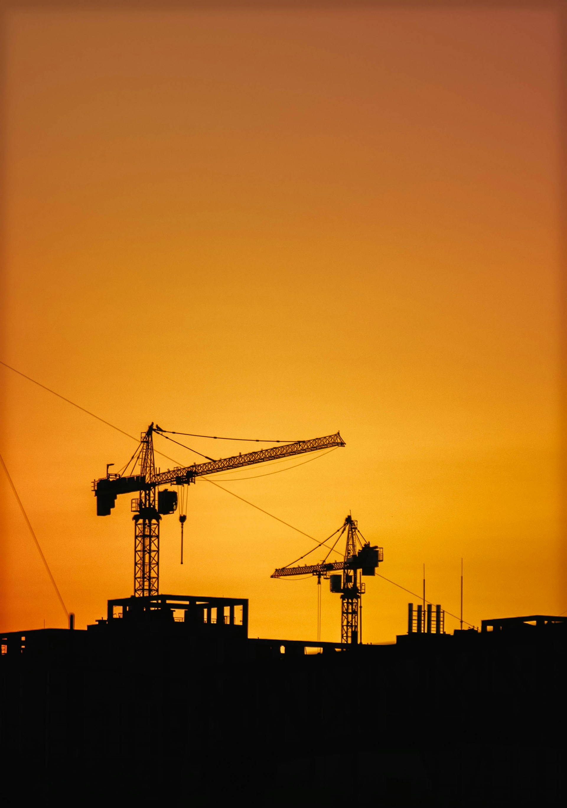 Construction cranes silhouetted against an orange sunset sky.
