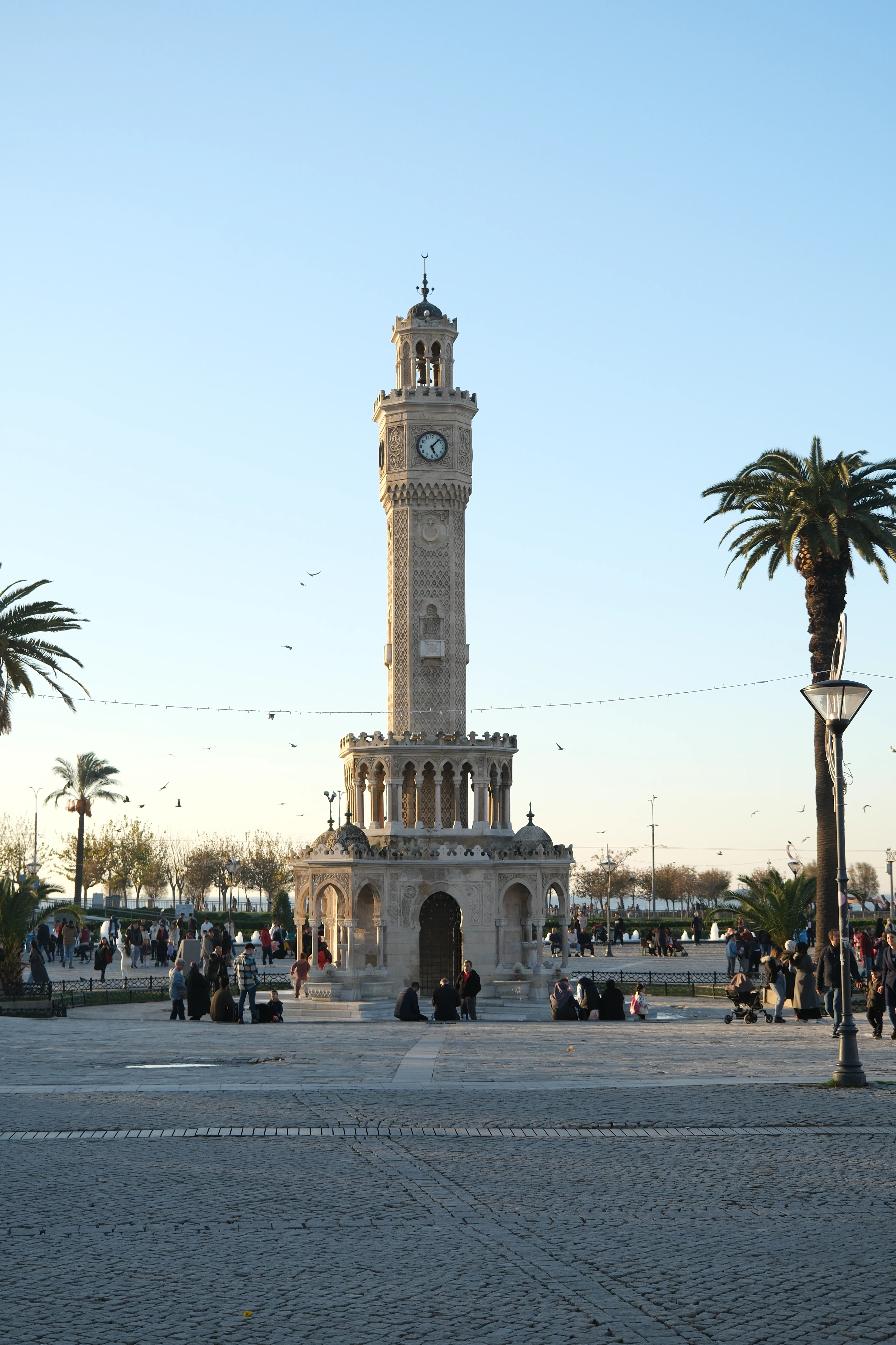 The Ottoman-era Izmir Clock Tower rising over Konak Square, its carved stone pavilion ringed by people sitting on the steps, with date palms and streetlamps around the plaza