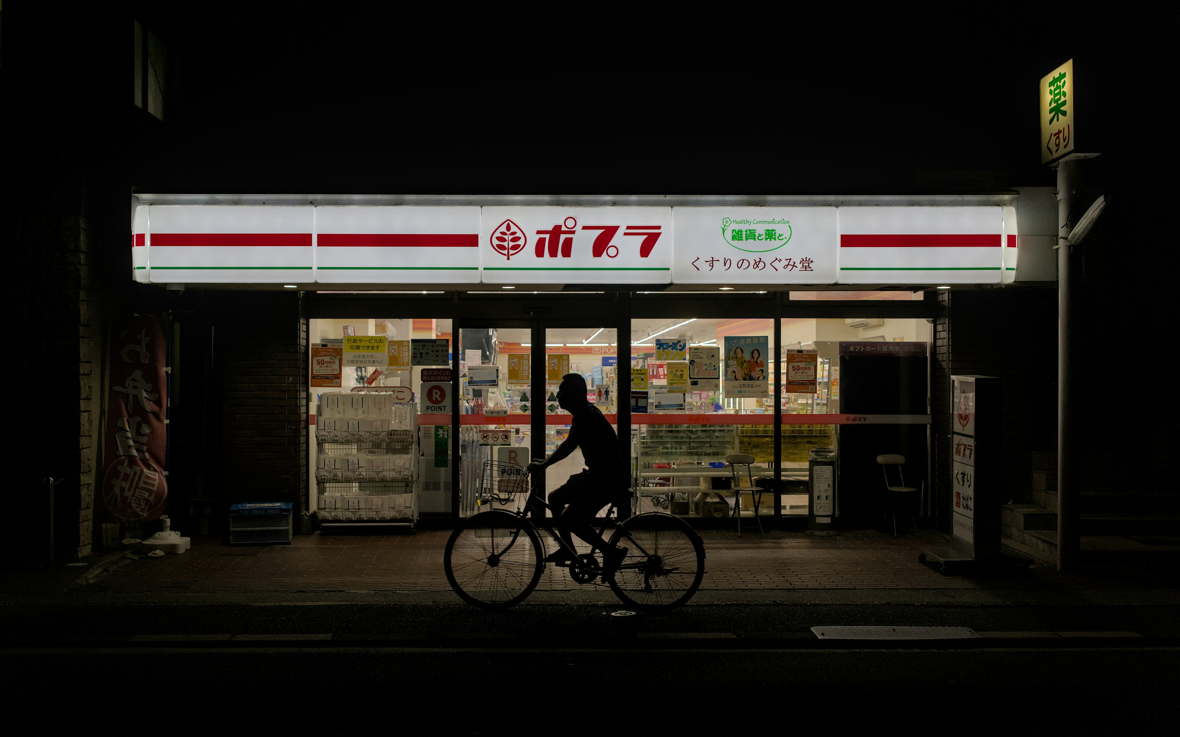 Inside a brightly lit Japanese convenience store (konbini) at night, showing shelves of instant noodles and ready meals, with a focus on a hot food counter