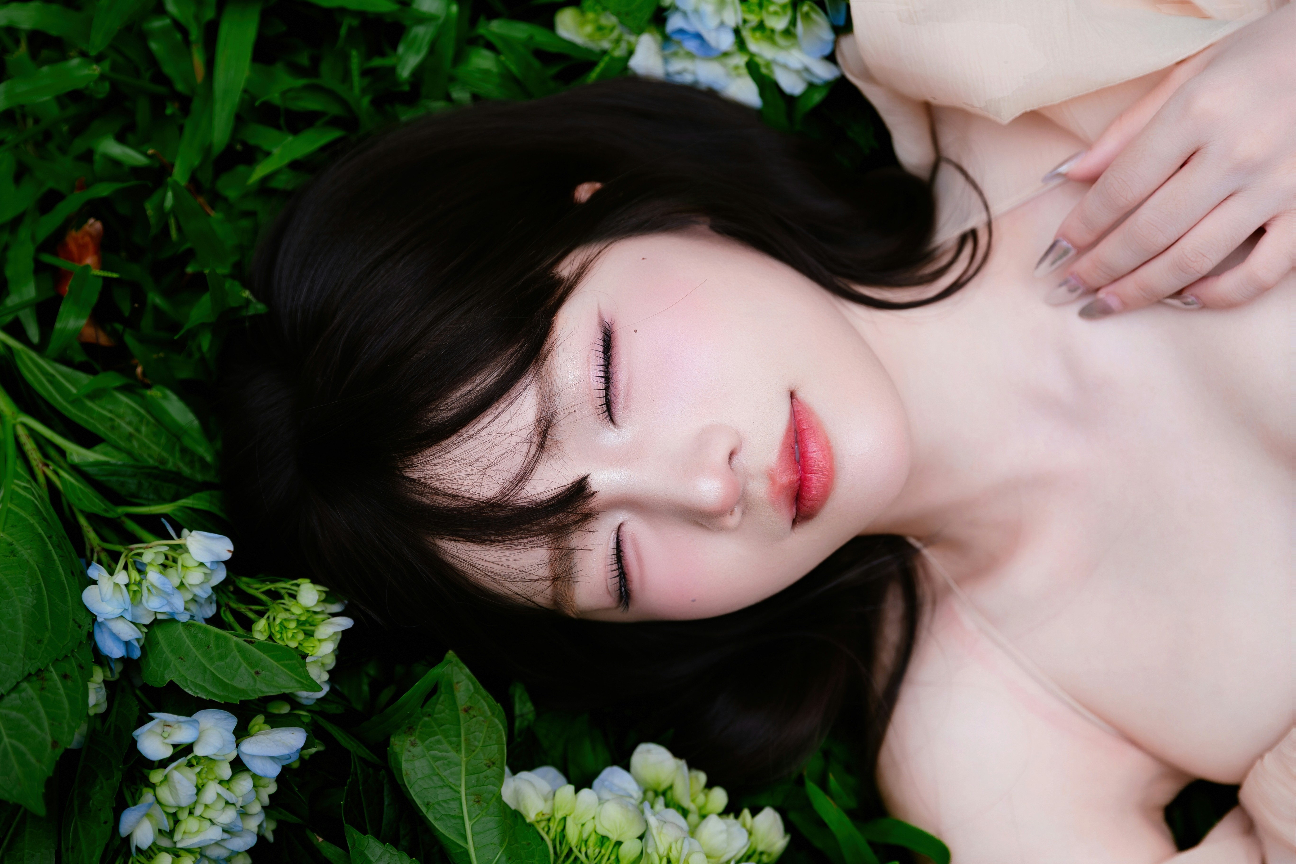 Smiling young woman lying on green grass with a bouquet of fresh hydrangea flowers, wearing a soft cream dress in natural light.