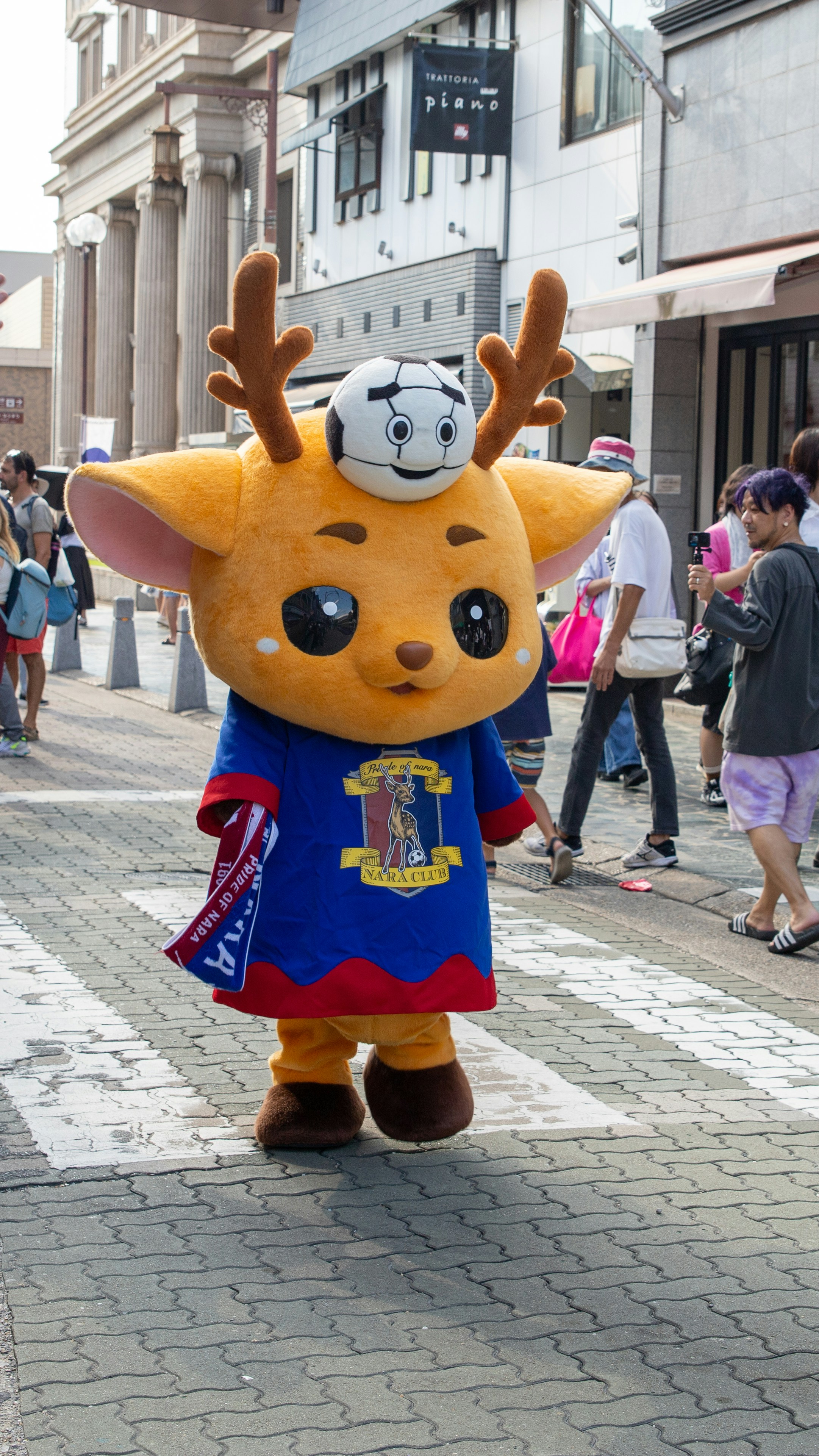 Mascot with a soccer ball on its head crosses street.
