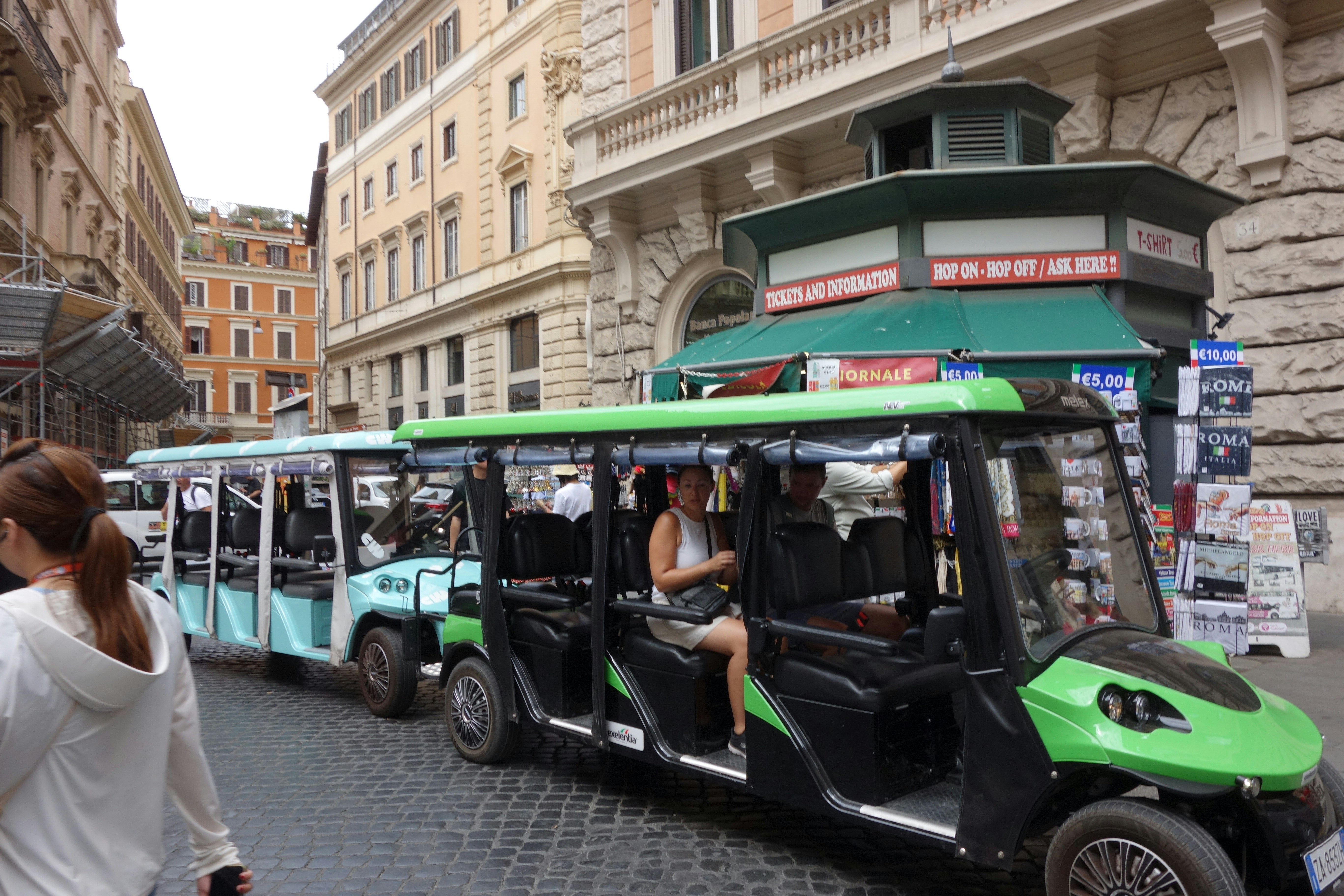 Tourists ride in open-air electric vehicles on cobblestone street.
