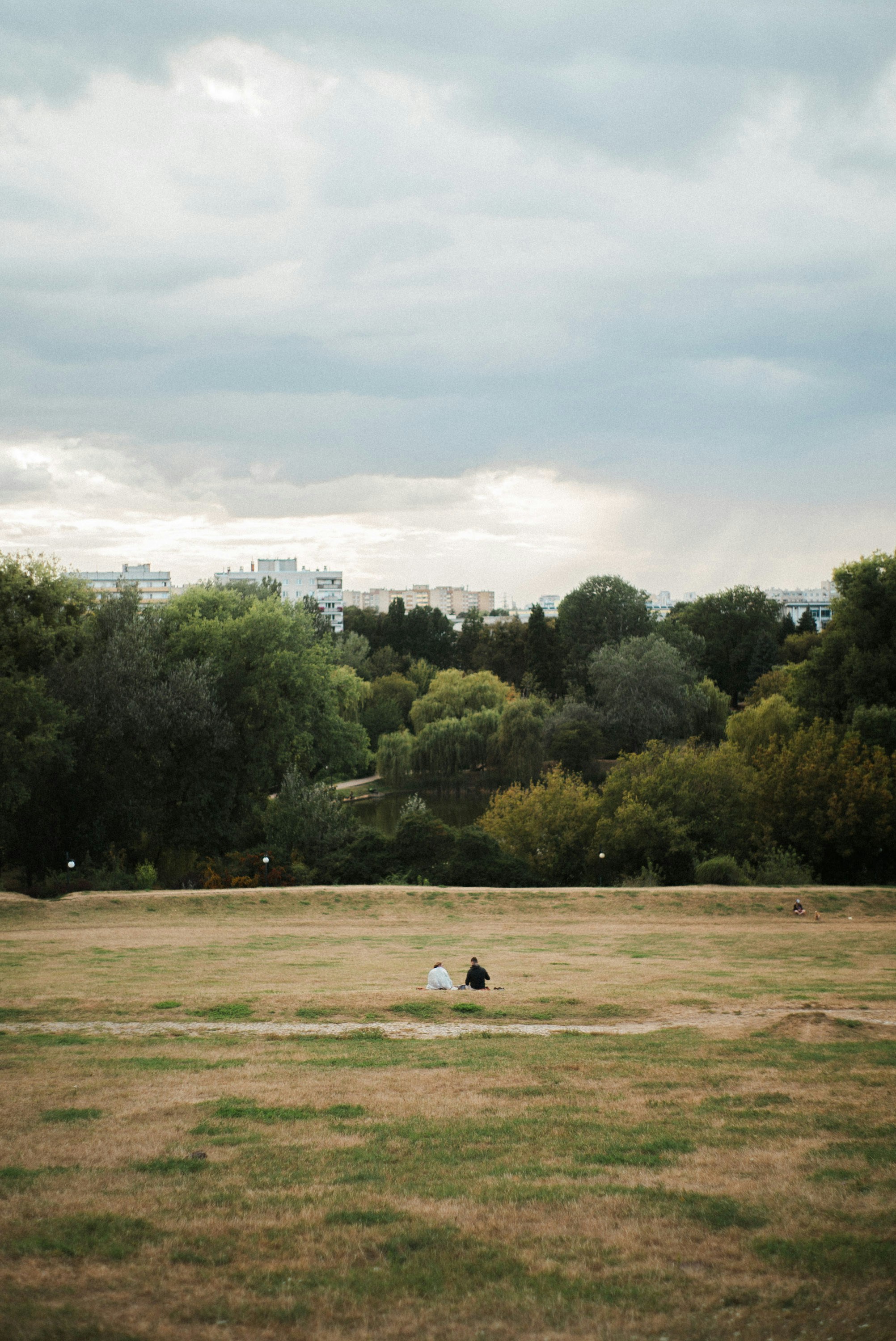 Two people sit on a grassy field with trees behind.