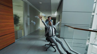Man in suit rides office chair pushed by colleague